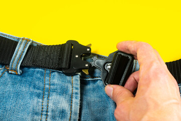 A man buttoned a black cloth belt on jeans. Modern men's waistband with dark matted metal buckle.