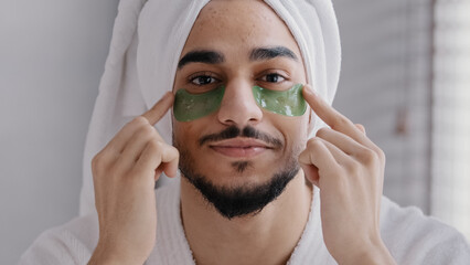 Front view portrait male face smiling indian arabian man wearing bath towel head glue sticky hydrogel eye-patches cosmetic procedures skin care guy looking at camera with collagen patches under eyes
