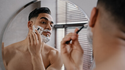 Arab Indian bearded man naked guy looks at reflection in mirror applies white foam shaving gel male cosmetics to beard shaves with disposable razor prepares for date at work day in bath at morning