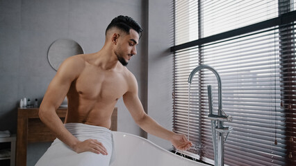 Millennial bare naked muscular indian arab man guy with beard wrapped towel around hips sitting on bathrub in bathroom at home turning faucet tasting hot cold water with hand getting ready for bathing