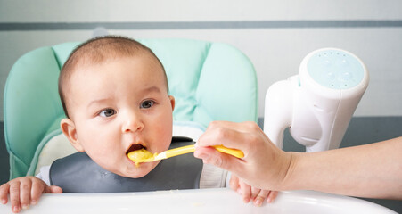 Mom feeds the baby with a spoon of vegetable puree at the children's feeding table. Baby's...