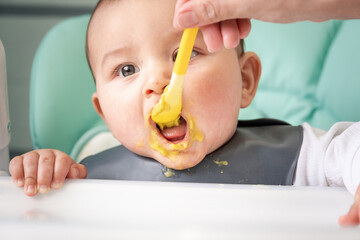 Mom feeds the baby with a spoon of vegetable puree at the children's feeding table. Baby's...