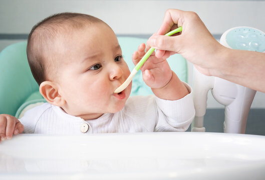 Mom feeds the baby with a spoon of vegetable puree at the children's feeding table. Baby's appetite, healthy nutrition, introduction of complementary foods. Copyspace, mock up