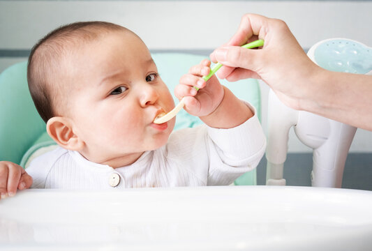 Mom feeds the baby with a spoon of vegetable puree at the children's feeding table. Baby's appetite, healthy nutrition, introduction of complementary foods. Copyspace, mock up