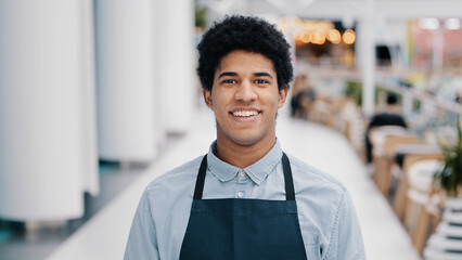 Male portrait smiling happy friendly African cafe pub worker biracial professional man waiter in apron uniform small business owner posing indoor looking at camera toothy face expression in restaurant - Powered by Adobe