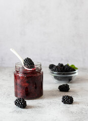 Naklejka na ściany i meble Blackberry jam in a transparent jar on a light table. Ripe blackberries in a glass bowl in the background. Light background