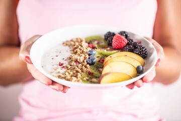 Yogurt, blackberry muesli, raspberries, blueberries, kiwi and peaches in a white bowl holding young woman
