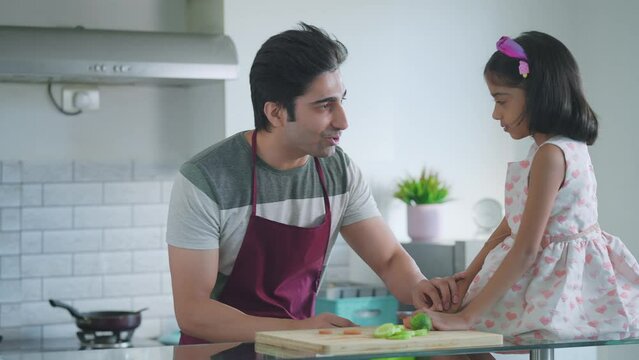 A young modern happy Indian Asian male or man wearing a chef apron is interacting and preparing a meal with a female kid or a girl in the kitchen. family, relationship Father or daughters day concept
