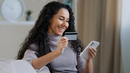 Smiling young hispanic woman hold phone and credit card happy girl customer shopping in online store pay bills using E-banking app service female rejoices successful purchase modern technology concept