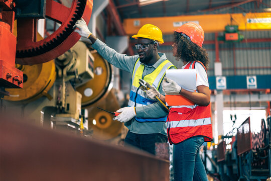 Two professional engineer/worker/technician use clipboard discuss work, walk in steel metal manufacture factory plant industry. Black African American man and woman wear hard hat check quality machine