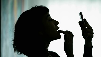 Young woman applying makeup. Girl removing lipstick looking at smartphone camera in silhouette
