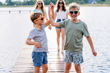 Four cute different age boys and girls sitting on a wooden platform by the river or lake dipping their feet in the water on warm summer day. Outdoor activities for kids concept.