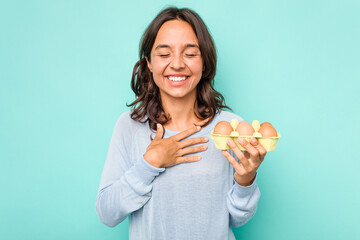 Young hispanic woman holding eggs isolated on blue background laughs out loudly keeping hand on...