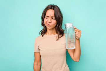 Young hispanic woman holding a water of jar isolated on blue background confused, feels doubtful and unsure.
