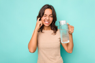 Young hispanic woman holding a water of jar isolated on blue background covering ears with hands.