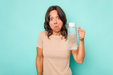 Young hispanic woman holding a water of jar isolated on blue background shrugs shoulders and open eyes confused.