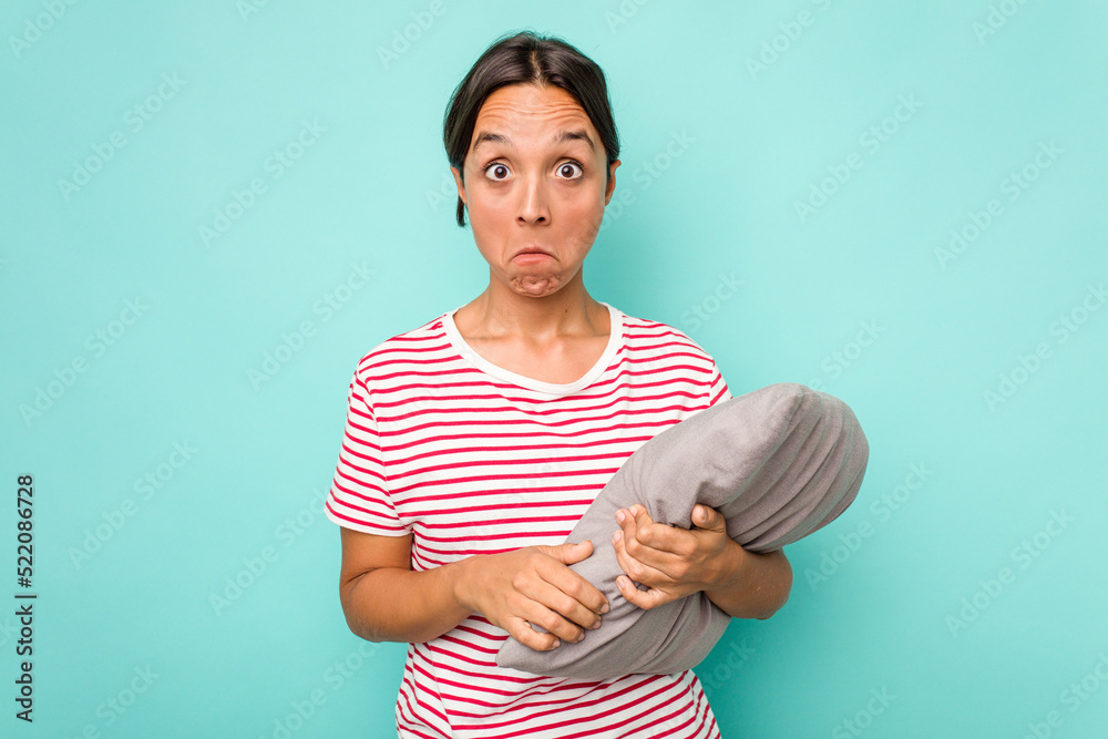 Wall mural Young hispanic woman holding a cushion isolated on white background shrugs shoulders and open eyes confused.