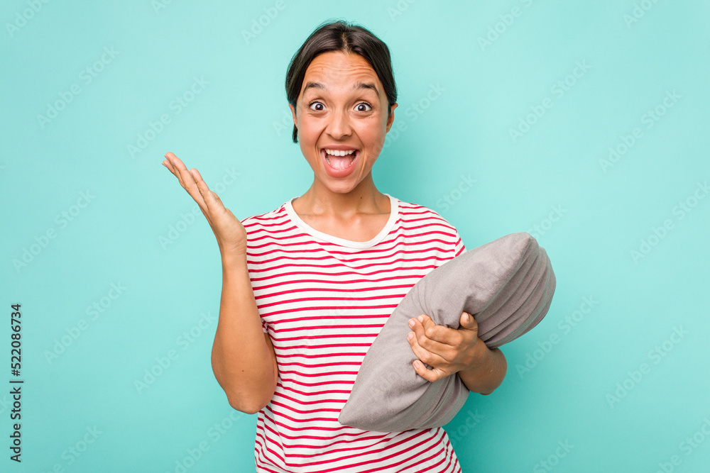 Wall mural Young hispanic woman holding a cushion isolated on white background receiving a pleasant surprise, excited and raising hands.