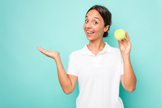 Young hispanic physiotherapy holding a tennis ball isolated on blue background showing a copy space on a palm and holding another hand on waist.