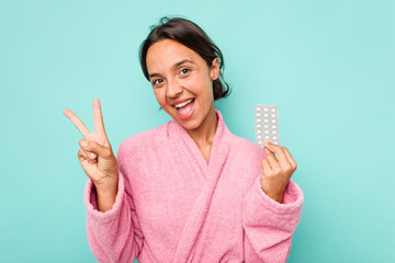 Young hispanic woman holding pills isolated on blue background joyful and carefree showing a peace symbol with fingers.