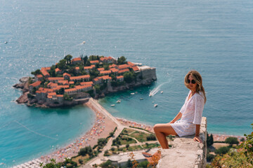 Fototapeta na wymiar a girl poses on a mountain with a top view of the city of Sveti Stefan in Montenegro, a picturesque bay