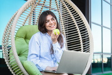 A young woman eats an apple and works on a laptop.