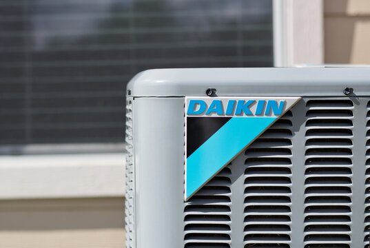 Daikin air conditioner unit, corner view at eye level with logo and domestic window in the background.