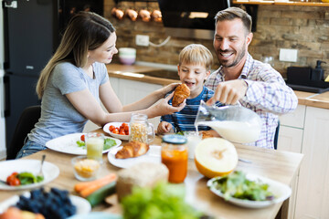 Young happy parents feeding their son with croissant during breakfast time in the kitchen