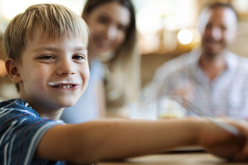 Happy little boy having yogurt mustache at home and looking at camera. There are people in the background