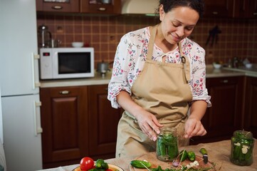 Attractive dark-haired multiethnic woman, housewife in beige chef's apron, makes pickled cucumbers at home kitchen. Homemade conservation of organic harvested seasonal vegetables. Pickling. Canning