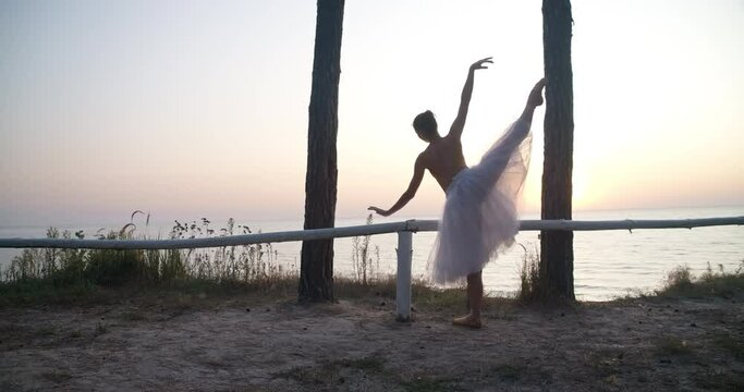 Graceful Caucasian ballerina dancing on tiptoes at sunrise on river bank. Back view wide shot of young slim talented woman performing in sunshine outdoors. Cinema 4k ProRes HQ