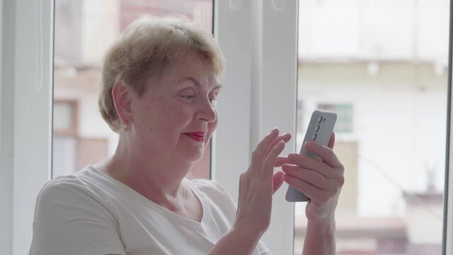 Older Middle Aged Blond Woman using and text messaging by mobile phone near Window with old town background, indoor, Relaxed mature old 60s woman, search ecommerce offers on cell phone technology devi