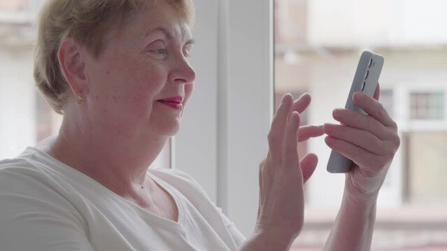 Older Middle Aged Blond Woman using and text messaging by mobile phone near Window with old town background, indoor, Relaxed mature old 60s woman, search ecommerce offers on cell phone technology devi