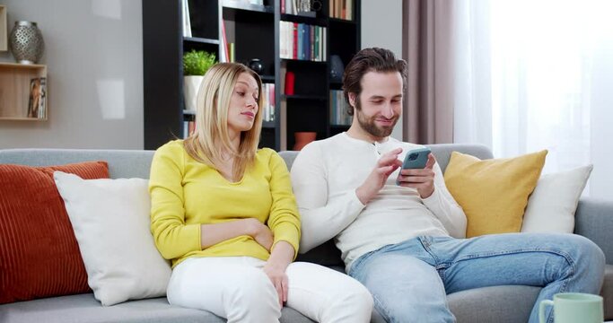 Caucasian handome man using smartphone while sitting on sofa at home. Beautiful woman watching her husband texting and chatting on phone. Female reaction on male chat online. Indoors.
