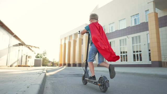 Child kid in superhero costume rides an electric scooter at school.Kid dream of travel and freedom.Schoolboy on an electric scooter in school yard.Child play in superhero costume and mask.Child winner