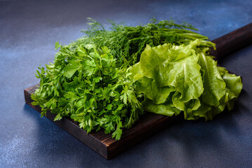 Salad, parsley and dill on a dark cutting board against a blue concrete background