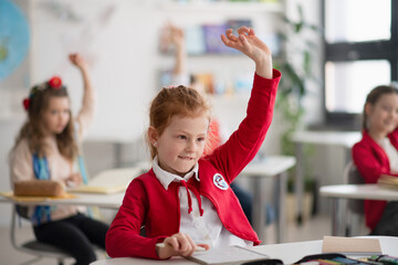 Happy schoolgirl with hands up at lesson in classroom at primary school.