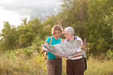 two middle aged women friends with backpacks watching the map standing on the mountain and hiking in nature and spending time together. travel tourism concept. Outdoor activities on weekends.