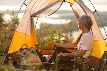 portrait of a smiling senior woman relaxed in a tent and drinks tea outdoors at sunset. copy space. Slow life. Enjoying the little things. spends time in nature in summer