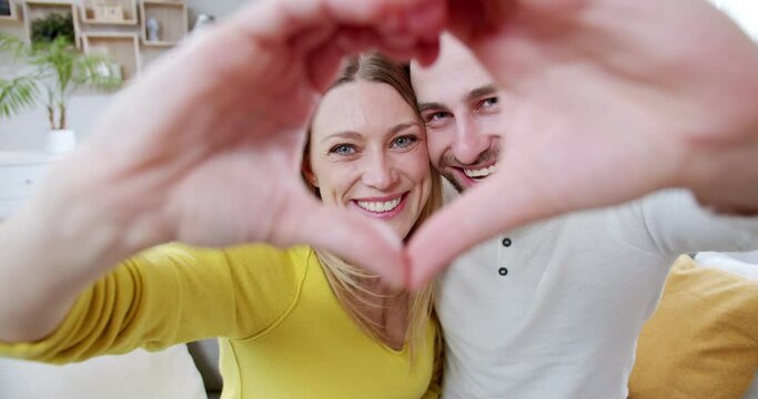 Camera zoming in and out on happy couple in love showing heart sign to camera and smiling while hugging. Portrait shot. Close up, joyful romantic man and woman demonstrating love. Feelings expression.
