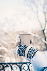 Hot drink in woman's hands wearing warm knitted gloves on cold winter snowy day.