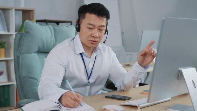 Busy businessman wear wireless headset making conference video call on computer. Skilled happy salesman advertising goods to customers, writing notes.