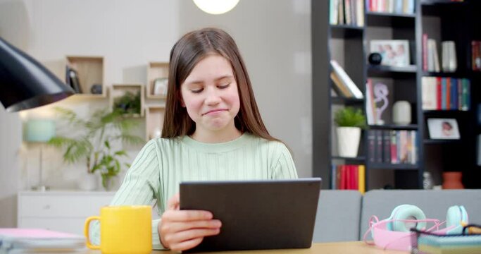 Cute Caucasian teen girl sitting at table in cozy living room and laughing while watching funny video on tablet device. At home. Beautiful happy teenage schoolgirl tapping and scrolling on computer.