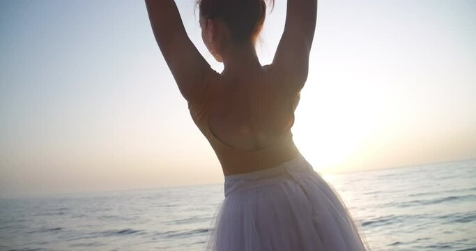 Back view slim woman in tutu dancing in sunbeam in slow motion on sea shore outdoors. Medium shot of graceful elegant ballerina performing in sunrays. Cinema 4k ProRes HQ