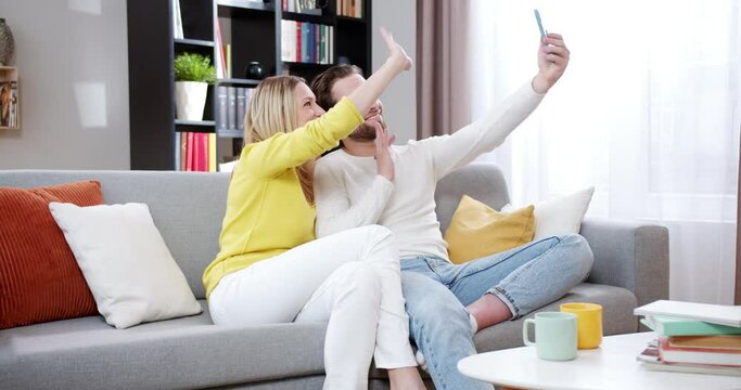 Cheerful Caucasian good-looking man and woman sitting on sofa in living room and videochatting on mobile phone. Happy married couple smling, waving with hands and having videochat on cellphone.