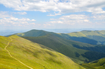Fototapeta premium Hiking trail through mountain ridge with green grassy slopes under blue sky with clouds on summer day. Carpathian Mountains, Ukraine
