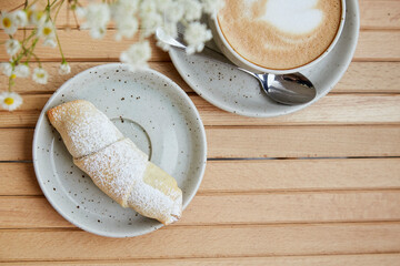 French homemade croissant crescent with powder. Cup of cappuccino in white ceramic cup, blurred foreground of chamomile flowers on wooden table at the terrace. Aesthetic breakfast. Copy space