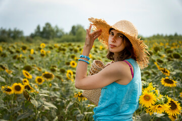 Young caucasian woman with straw hat and straw bag on sunflower field. Beautiful lady carrying bag with flowers among sunflower field. Pretty woman  smiles holding sunflower. Golden hour.