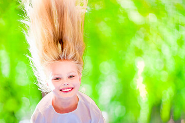 Cheerful young girl with long blond hair hangs upside down outdoors in the park. Sunny summer day