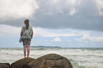 Young woman standing alone on the rocks looking at the beach. Cloudy day. Bombinhas, Santa Catarina State, Brazil